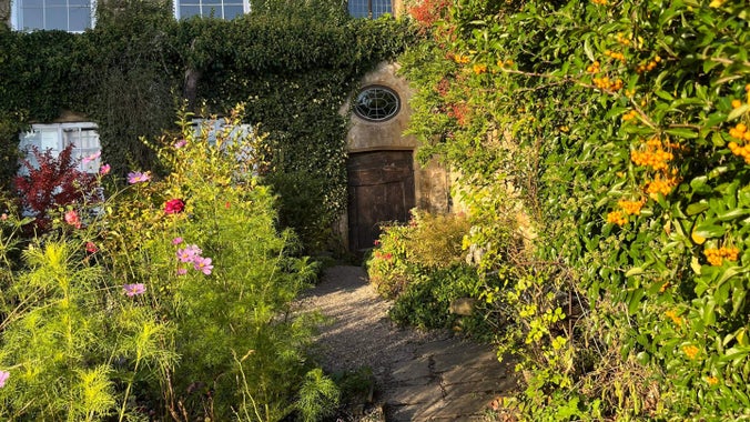 A path bordered by plants turning autumnal reds and oranges leads to an old wooden door to Crook Hall.